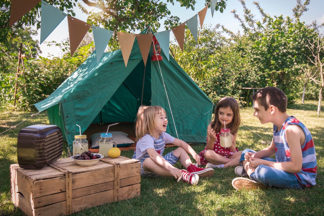 Three children sit on grass in front of a green tent, laughing and talking. A crate holds a radio, drinks, and snacks. Bunting hangs overhead, and the scene is set outdoors on a sunny day with trees and greenery.