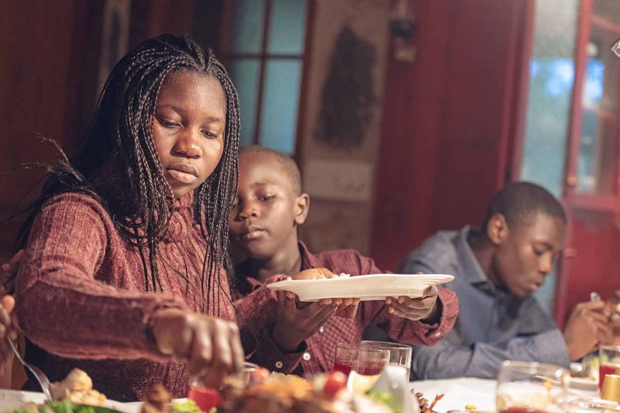 A young girl serves food at a dinner table while a boy next to her holds a plate and another looks down. The warm, cozy setting suggests a family meal, perhaps reflecting on Thanksgiving dinner cost traditions through the years.