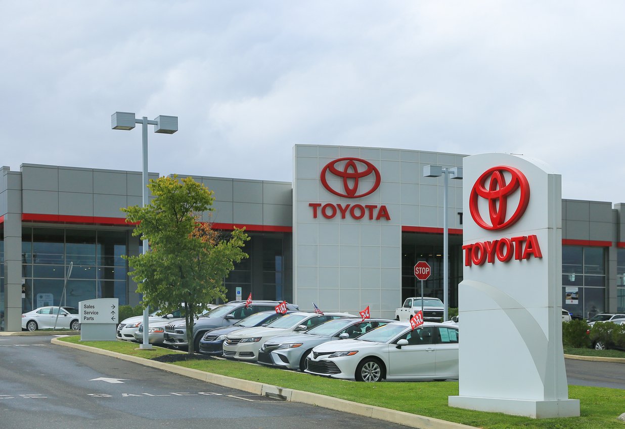 A Toyota dealership with several new cars parked outside, a large Toyota sign in the foreground, and a modern building with Toyota logos visible on a cloudy day.