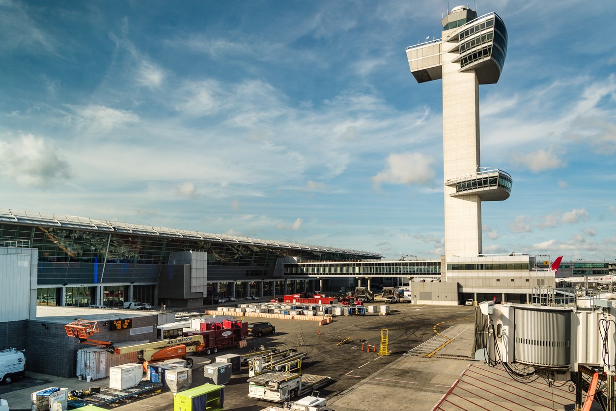 Modern airport scene with a tall air traffic control tower, terminal building, various ground vehicles, cargo containers, and clear blue sky with scattered clouds in the background.
