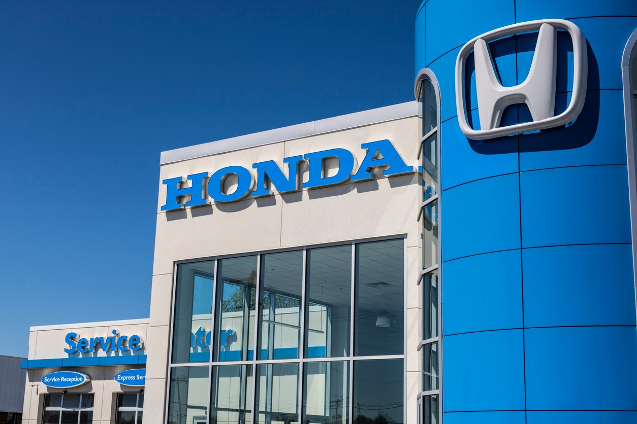 The exterior of a Honda dealership is shown, featuring a blue building with a large Honda logo and sign. A smaller building with "Service Center" signage is visible to the left, all under a clear blue sky.
