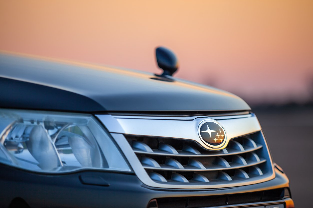 Close-up of the front grille and headlight of a dark SUV with a Subaru badge, photographed at sunset with a blurred background and a warm orange sky.