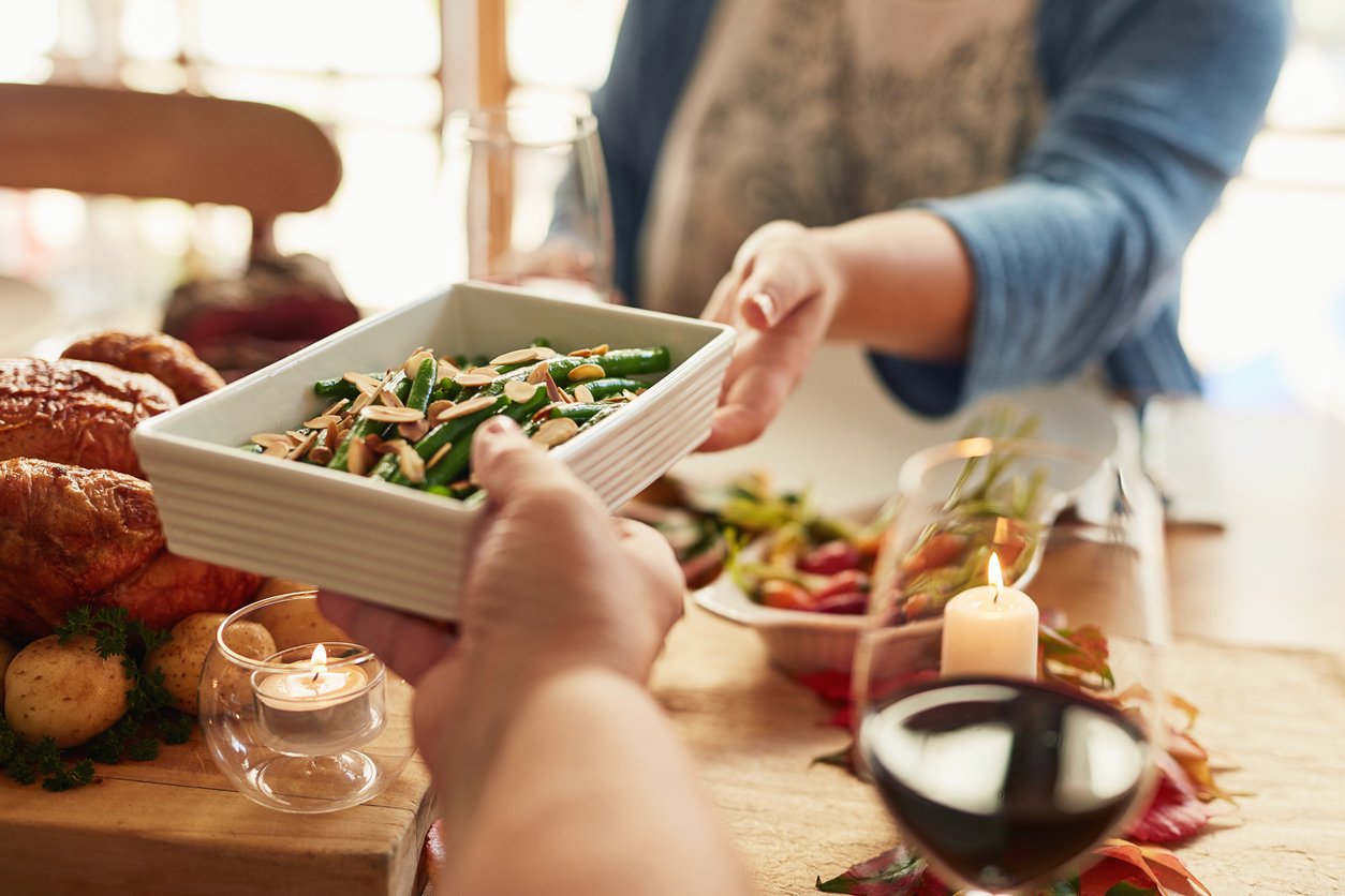 Two people pass a rectangular white dish of green beans with almonds at a table set with candles, wine, roasted chicken, potatoes, and other dishes, suggesting a cozy shared meal.