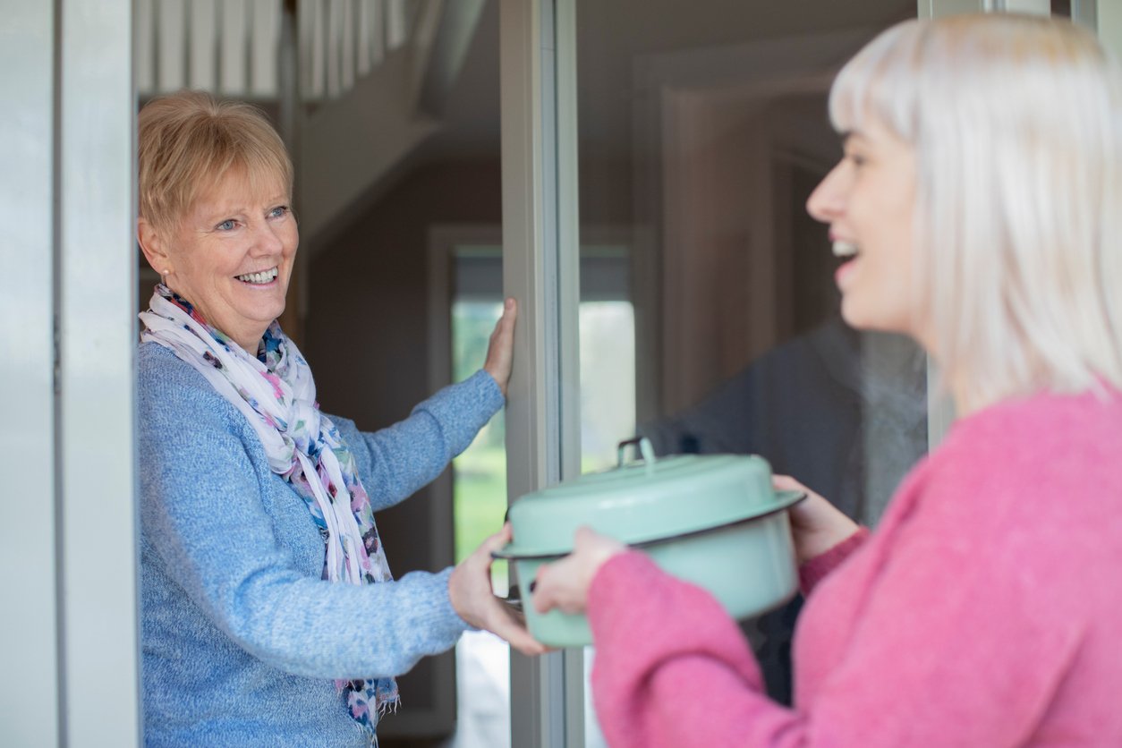 A smiling older woman opens her front door to greet a younger woman who is handing her a covered pot, suggesting a friendly visit or meal delivery.