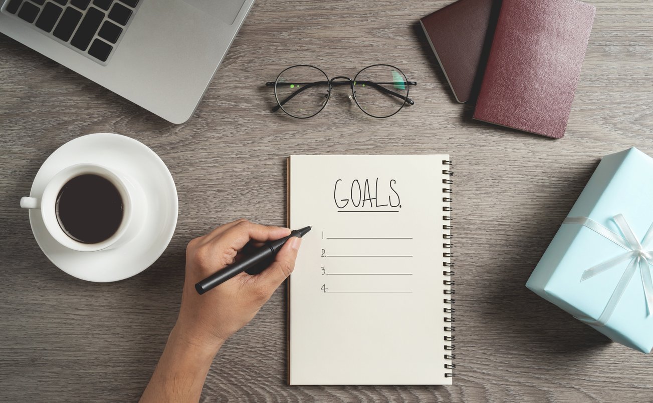 A hand writes in a notebook labeled "GOALS" with a numbered list. Surrounding items include a cup of coffee, eyeglasses, a laptop, two notebooks, and a blue gift box with a white ribbon on a wooden table.