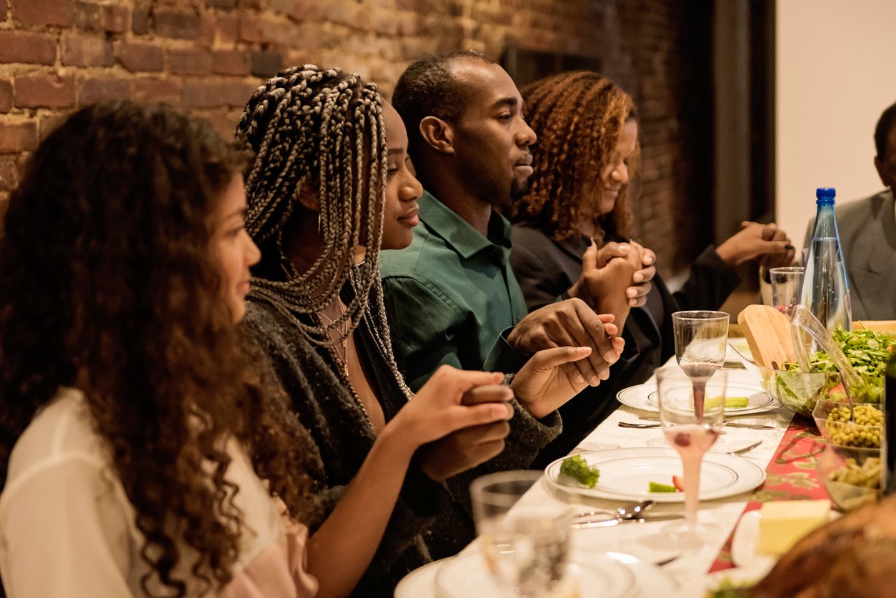Four people sit at a dinner table holding hands with eyes closed, appearing to pray before a meal. The table, reminiscent of Thanksgiving Dinner Cost History, is set with plates, glasses, salad, and a water bottle against a brick wall backdrop.
