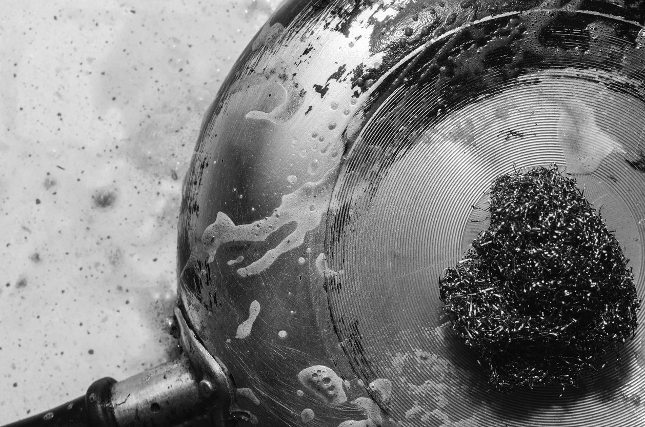 A close-up, black-and-white image of a soapy metal pan being scrubbed with a metallic scouring pad, with soap suds visible on the pan and the surface below.