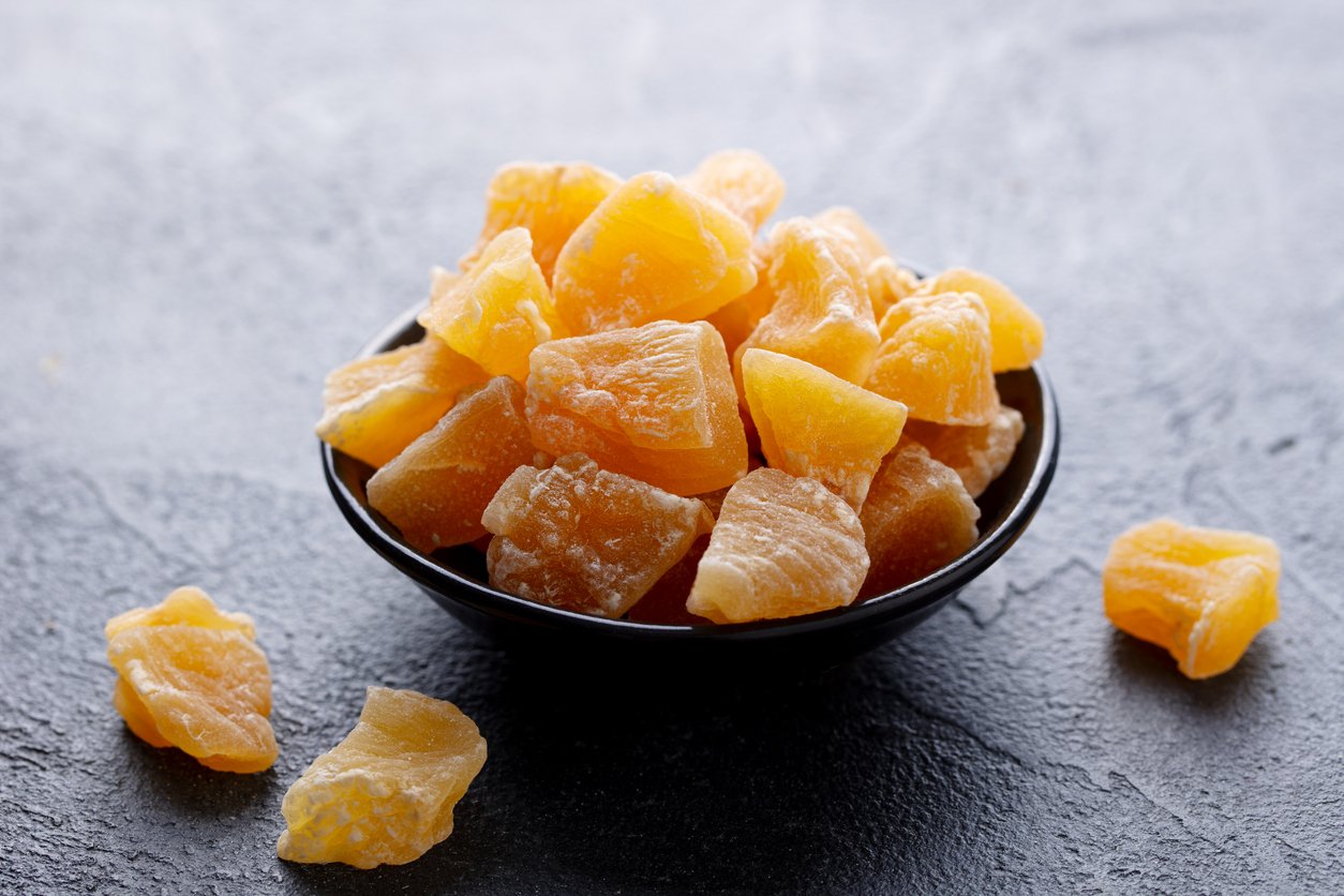 A small black bowl filled with bright yellow, cubed pieces of dried pineapple sits on a dark, textured surface. A few pineapple pieces are scattered outside the bowl.