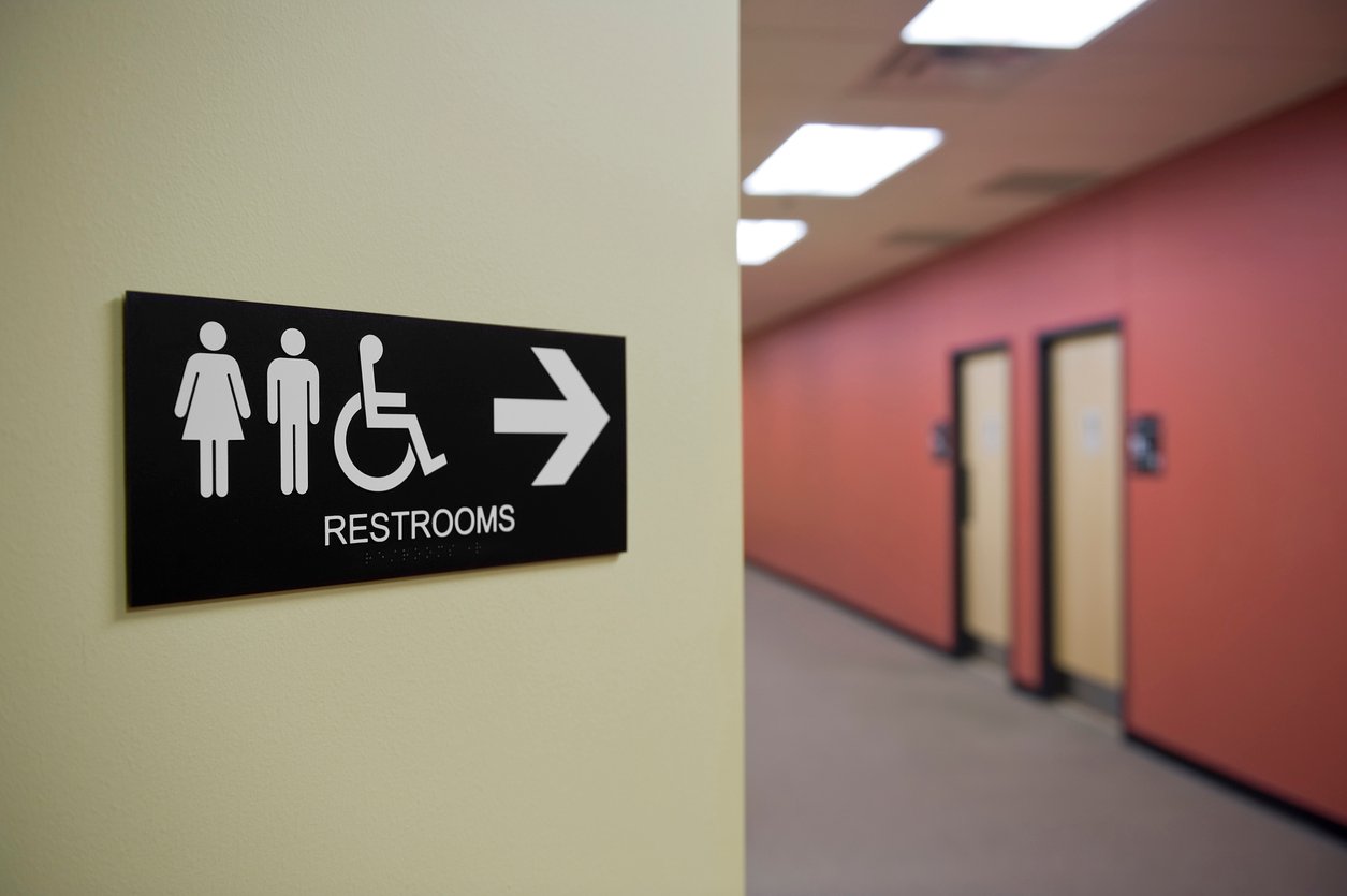 A black and white restroom sign with icons for women, men, and wheelchair access points right toward a hallway with restroom doors.