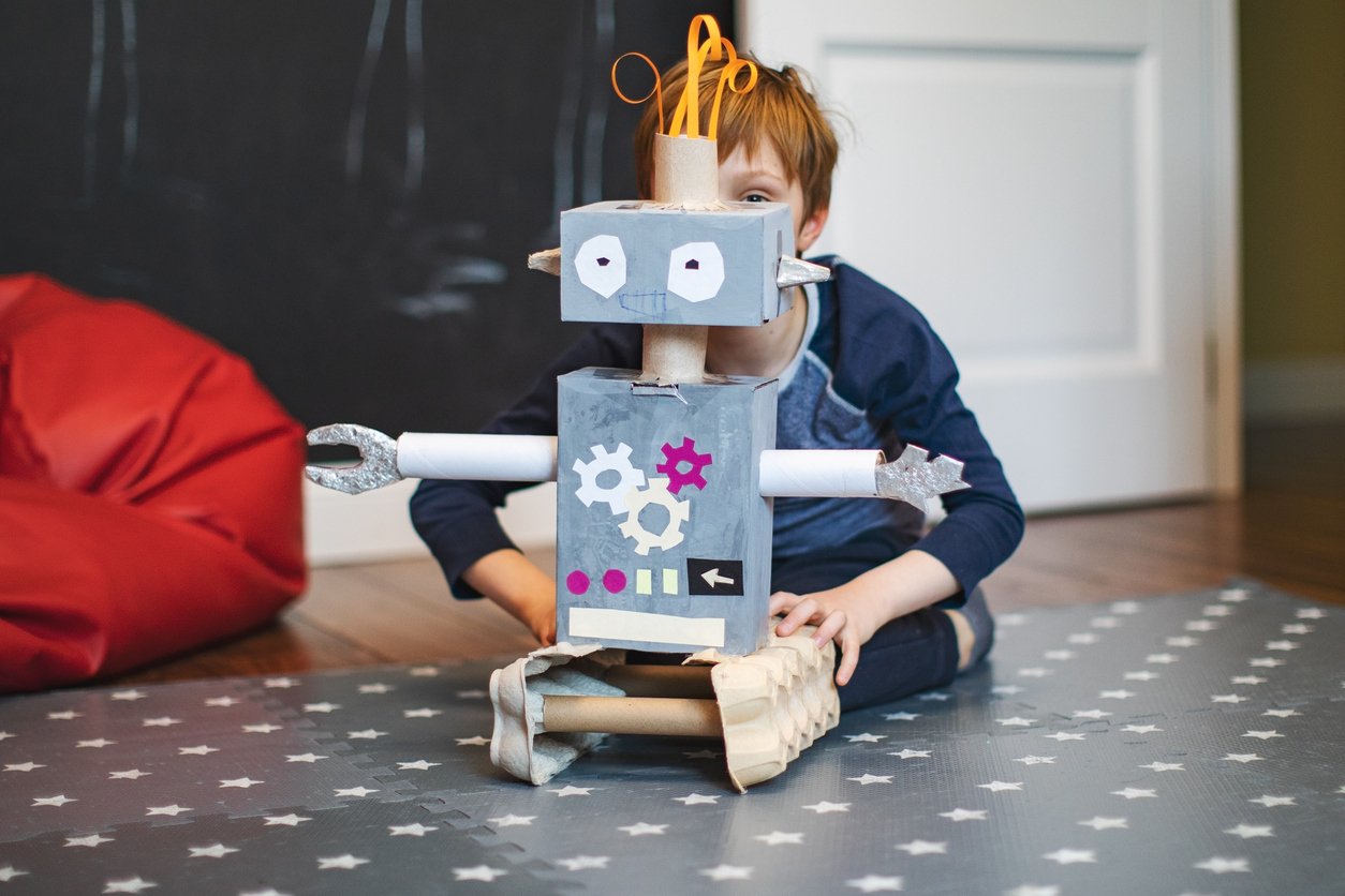 A child sits on the floor behind a homemade robot costume made from cardboard boxes, tubes, and colored paper, with gears and buttons drawn on the front, in a room with a star-patterned mat.