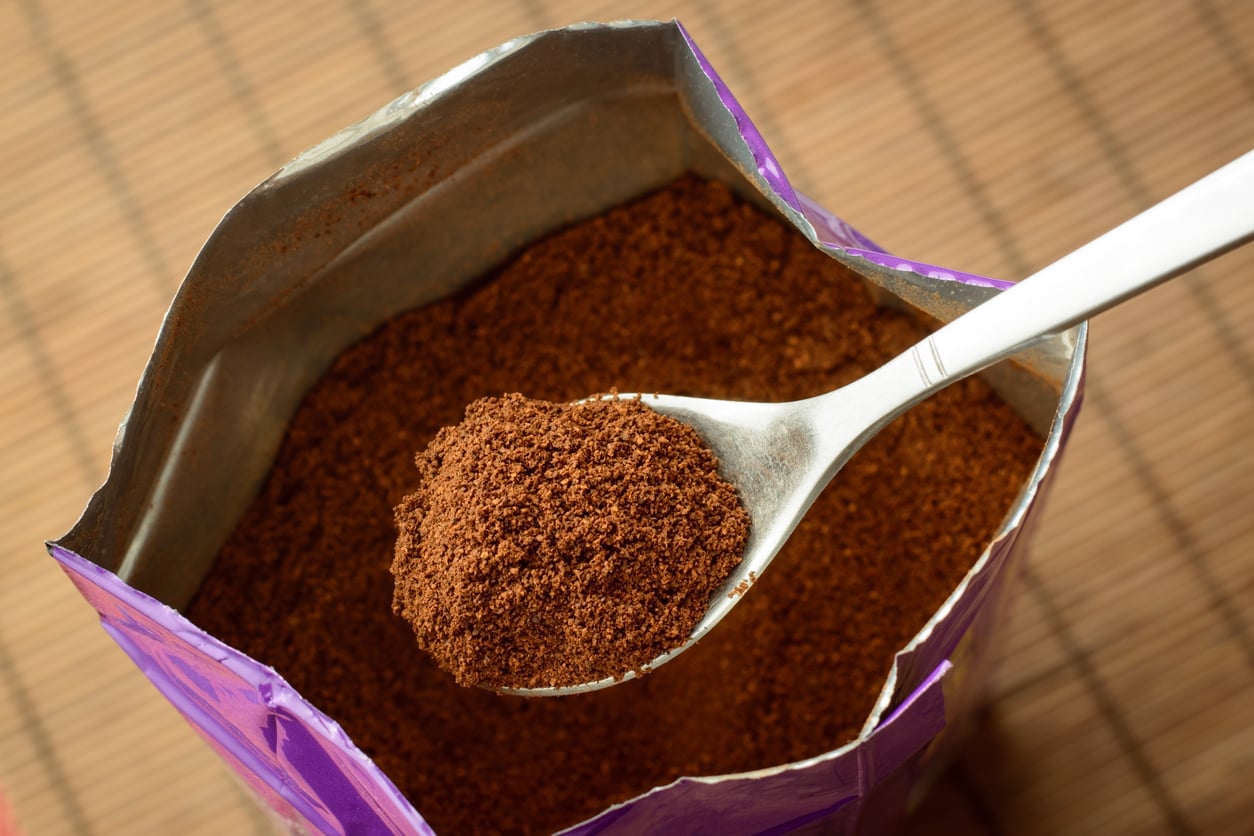 A close-up of a spoonful of ground coffee being held above an open bag filled with more ground coffee, with a bamboo mat surface in the background.