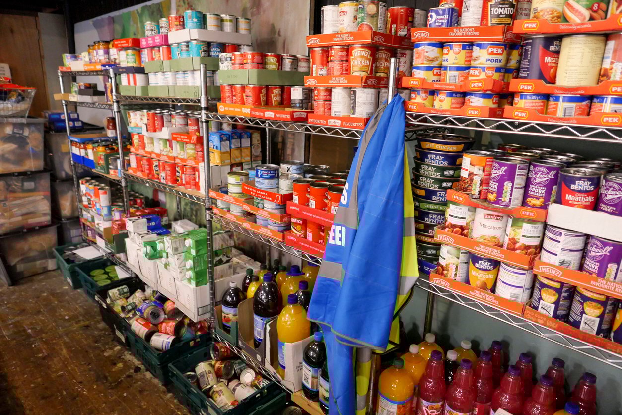 Shelves filled with canned goods, bottles of juice, and other non-perishable food items. A blue and yellow flag drapes over the lower shelf. The items are organized and stacked in a food pantry or storage room.