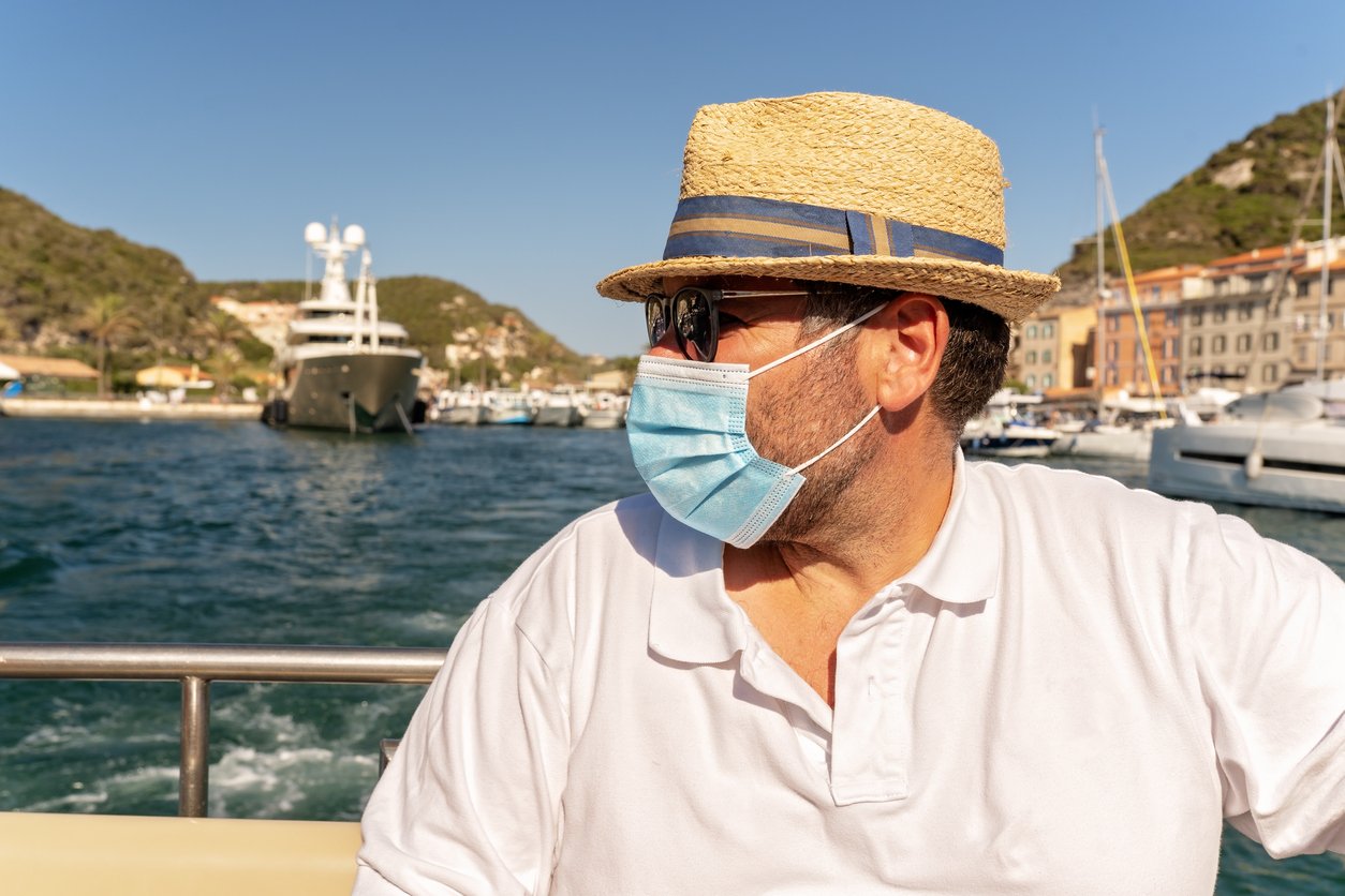 A man wearing a straw hat, sunglasses, and a face mask sits on a boat, looking to the side. Behind him are yachts, water, and buildings with green hills under a clear blue sky.