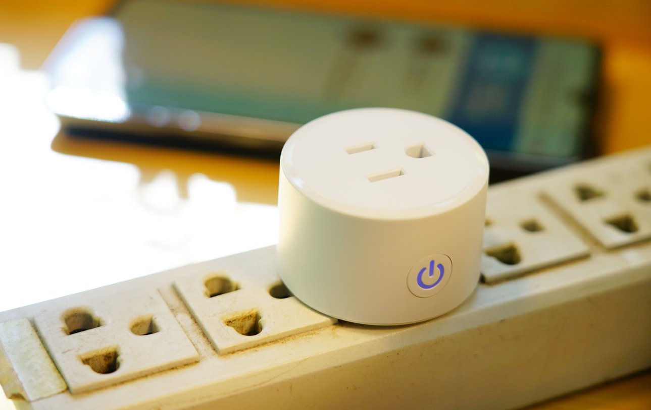 A small smart plug with a glowing power button sits on a white power strip, with a smartphone lying blurred in the background on a wooden surface.