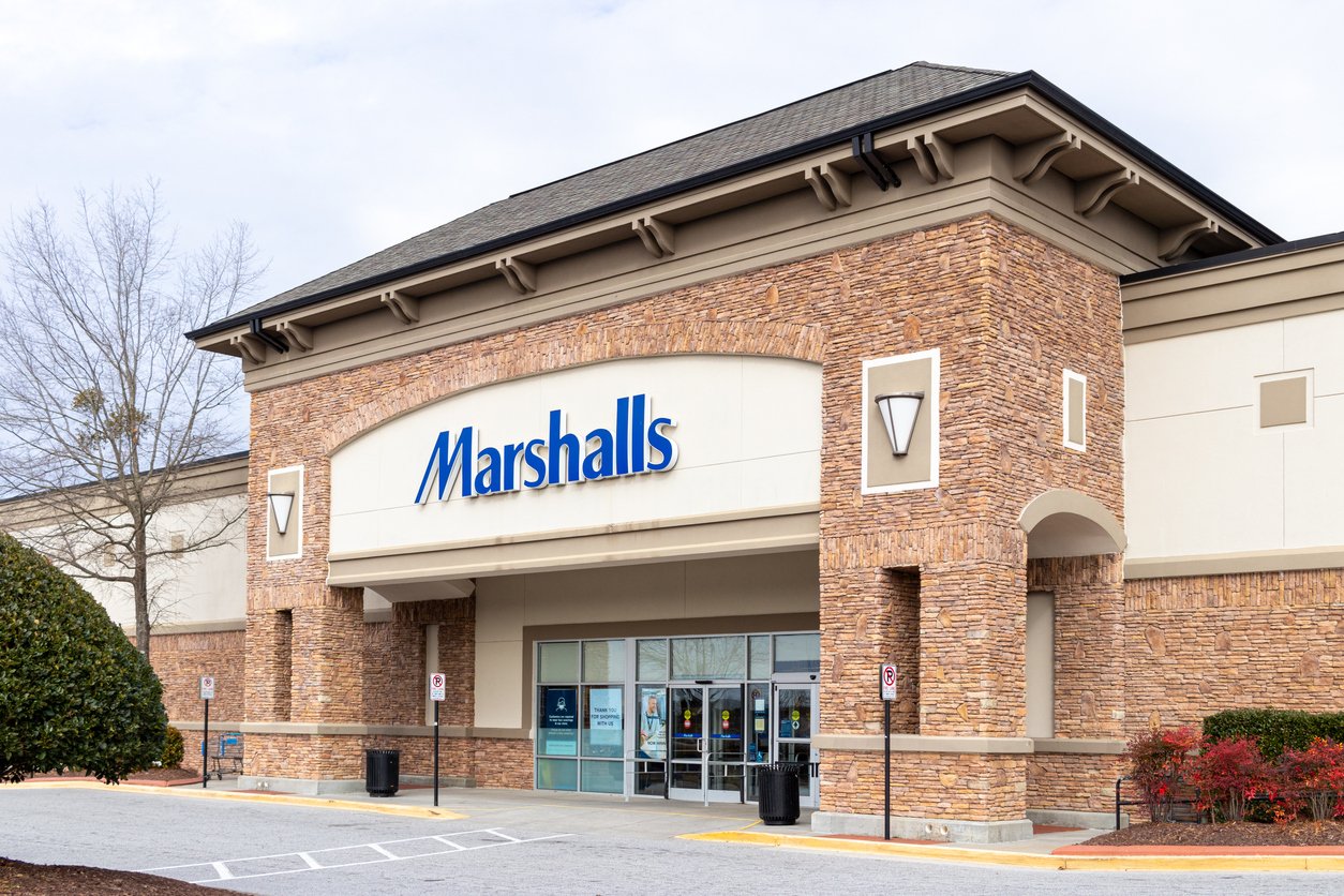 The entrance of a Marshalls store with a stone facade, large glass doors, and blue Marshalls sign, surrounded by bushes and a parking lot.
