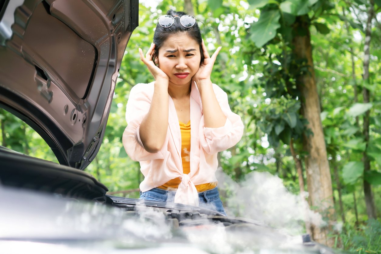 A woman stands in front of an open car hood in a wooded area, looking distressed and holding her head as steam rises from the engine.