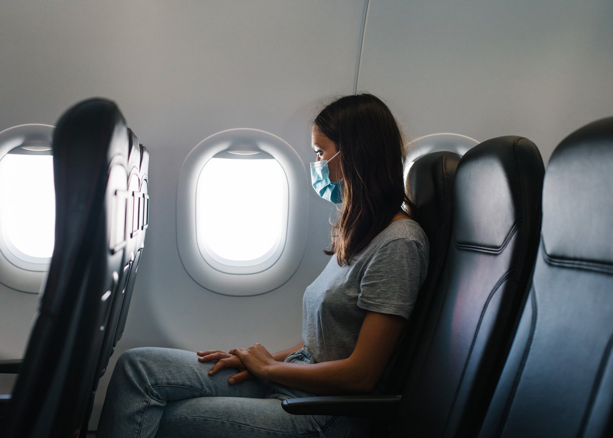 A woman wearing a face mask sits alone by the window on an airplane, looking outside. The cabin is well-lit, and the seats beside her are empty.