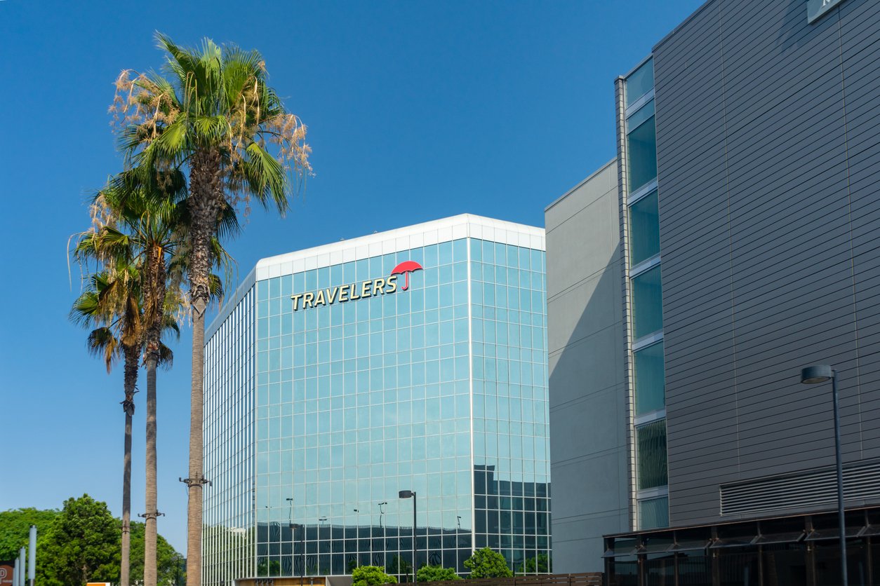 A modern glass office building with the "Travelers" logo and red umbrella on top, next to a tall palm tree and a neighboring metal-clad building, under a clear blue sky.