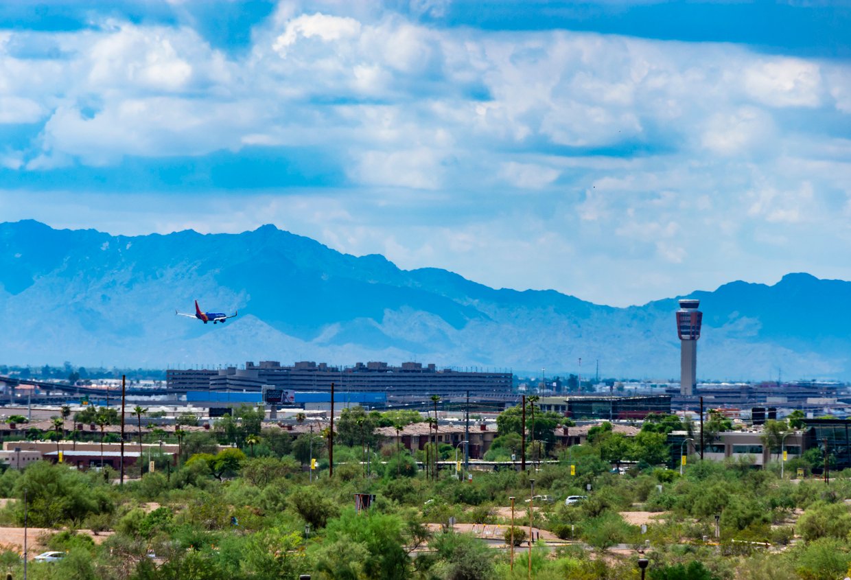 A commercial airplane is taking off near a city with a control tower, set against a backdrop of distant mountains under a partly cloudy sky. Green vegetation and buildings are visible in the foreground.