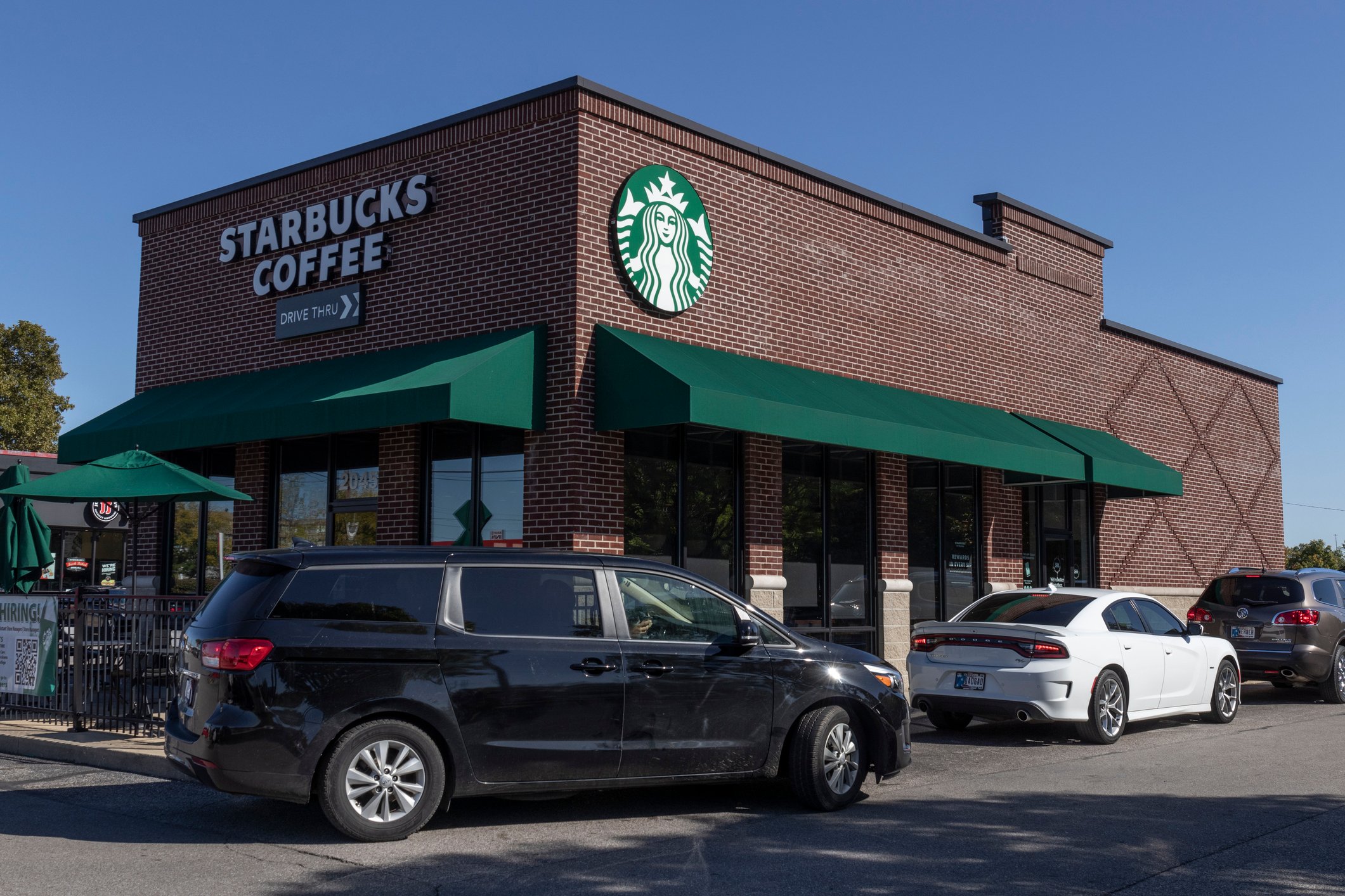 A Starbucks Coffee shop with green awnings is shown in a parking lot with several parked cars on a sunny day. The building is made of red brick and has the Starbucks logo displayed.