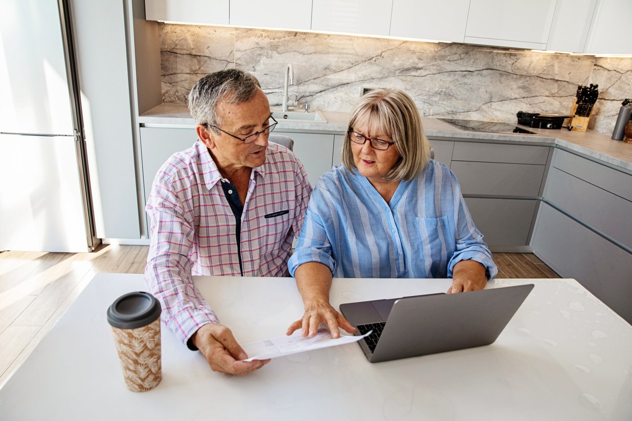 An older man and woman sit at a kitchen table with a laptop, discussing a document together. The man holds the paper while the woman gestures at the laptop. A coffee cup is on the table.