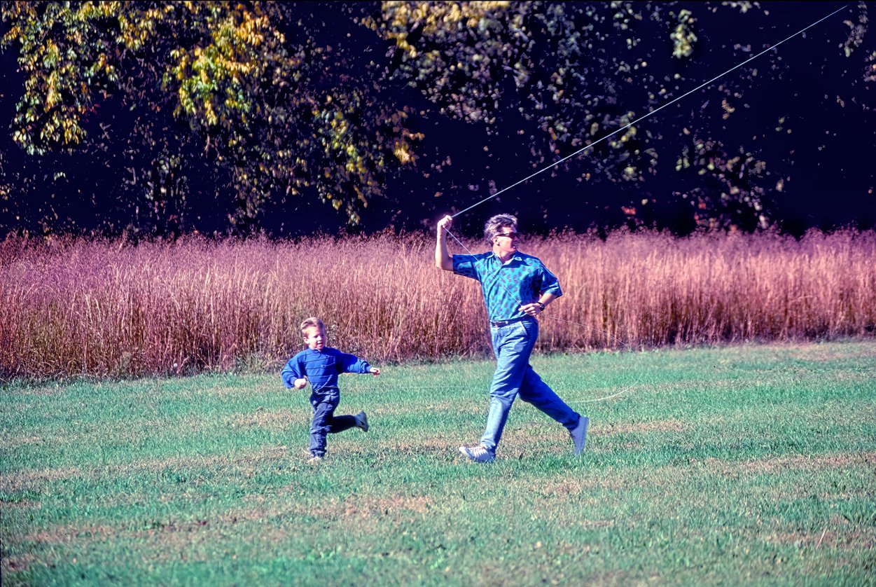 An adult and a child run through a grassy field, flying a kite together on a sunny day. Tall brown grass and trees with autumn foliage evoke the warmth of Thanksgiving Dinner Cost History in the background.