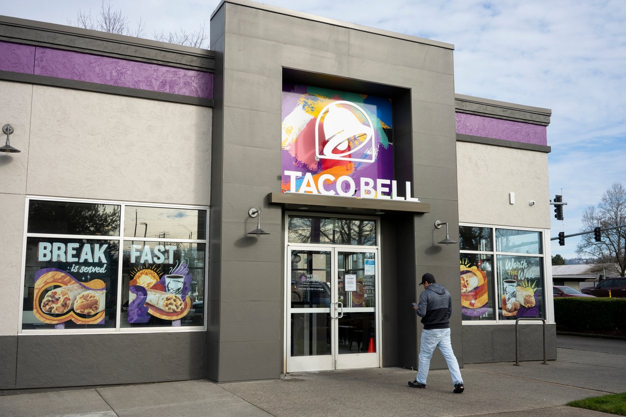 A person walks toward the entrance of a Taco Bell restaurant, with large colorful signs on the windows displaying breakfast menu items. The sky is partly cloudy.