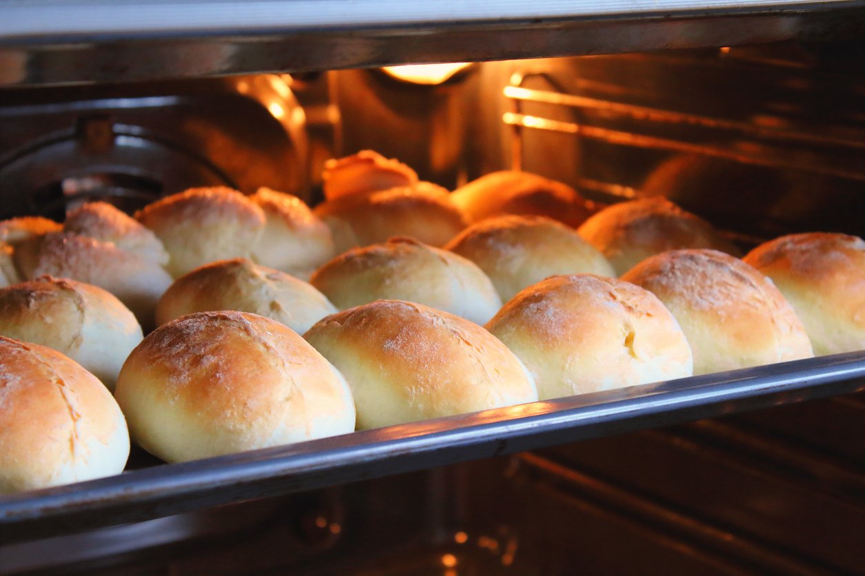 A tray of golden brown bread rolls is baking in an oven, with the rolls rising and getting lightly browned on top under the oven light.