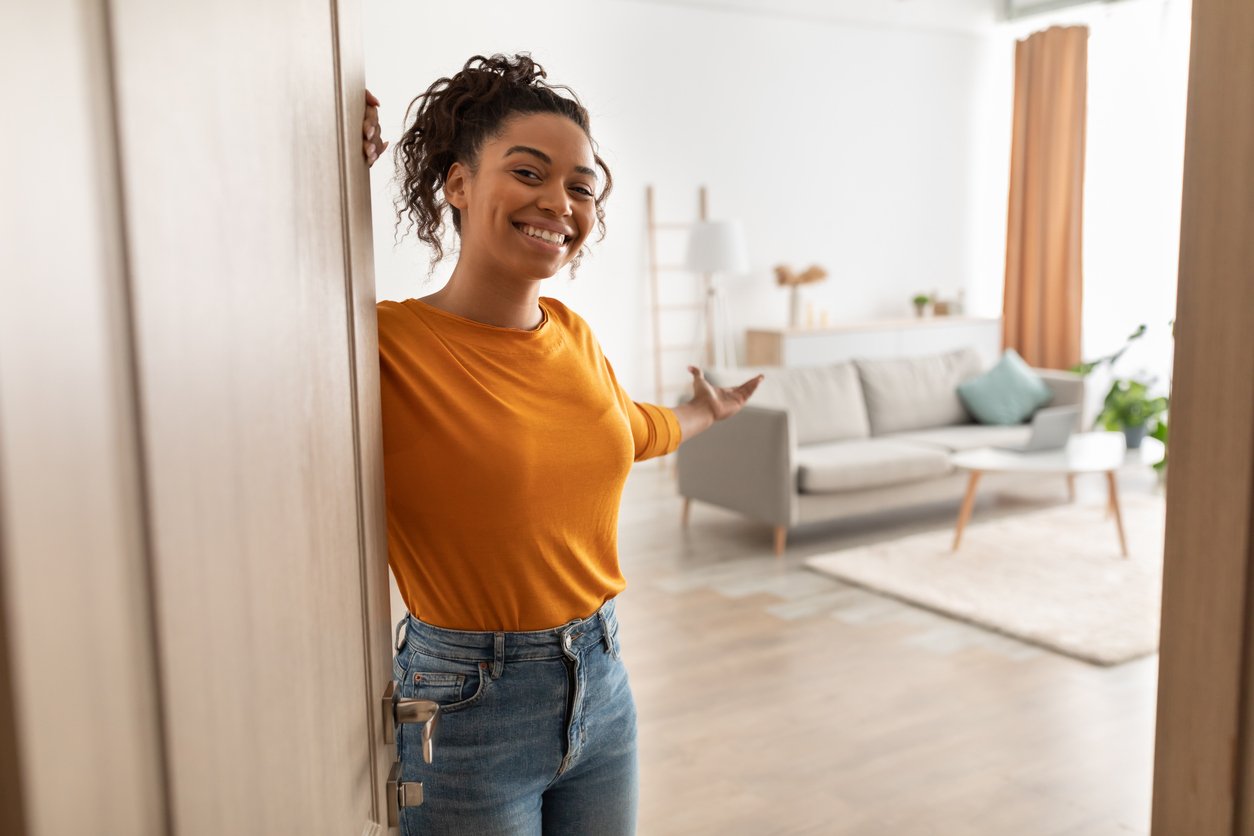A smiling woman in a yellow top and jeans stands at an open door, gesturing to invite someone into a bright, modern living room with a grey sofa and wooden decor.