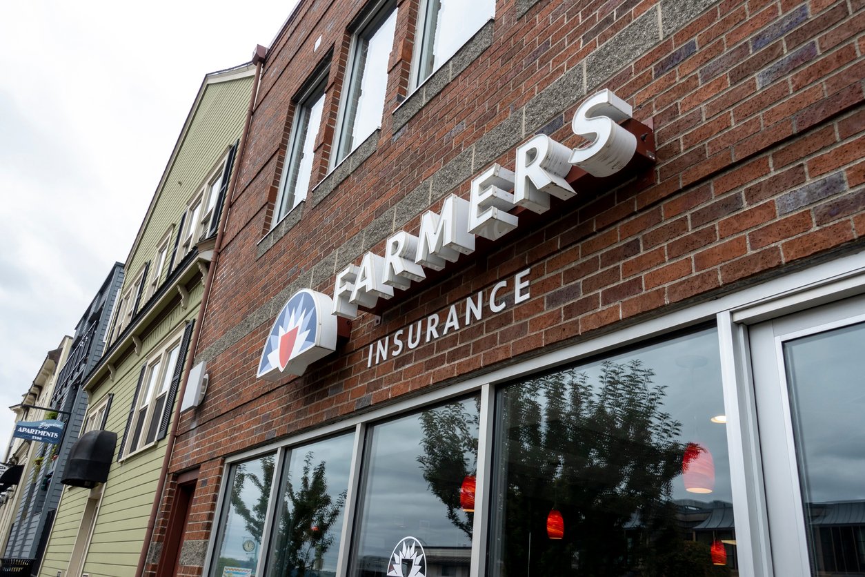 A brick building with large windows displays a "Farmers Insurance" sign above the entrance. Reflections of trees and red hanging lights are visible in the glass. Other buildings are visible along the street.