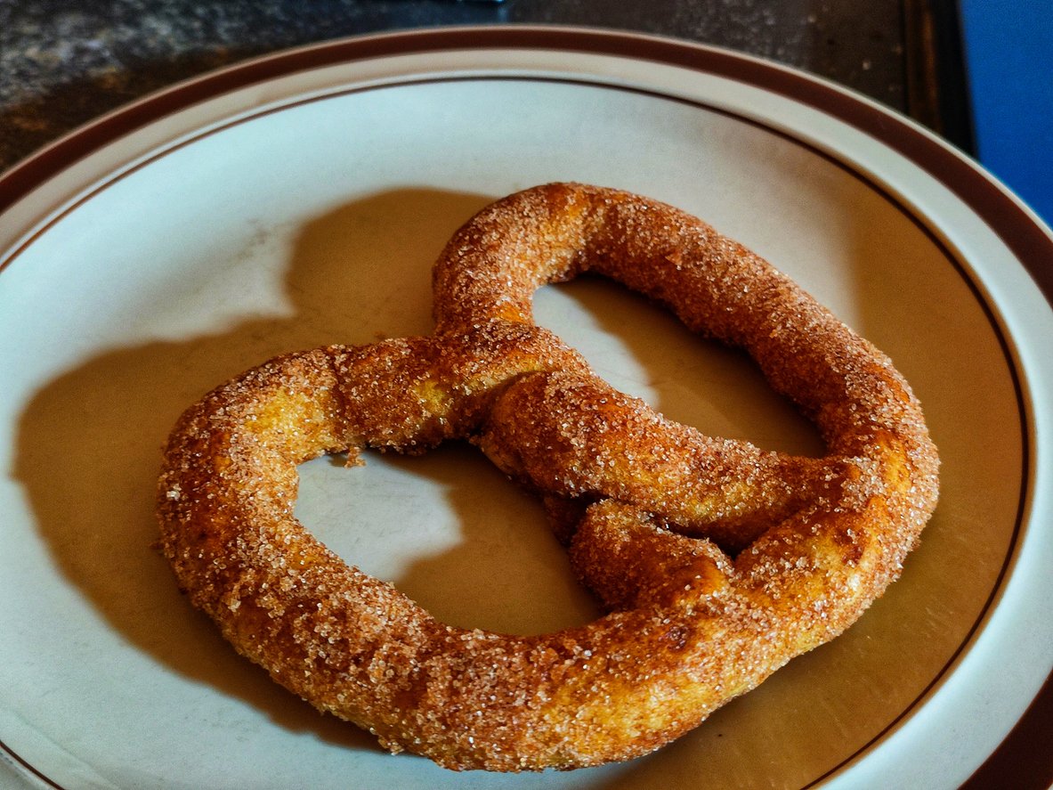 A cinnamon sugar pretzel sits on a white plate with brown trim, casting a soft shadow on the plate.