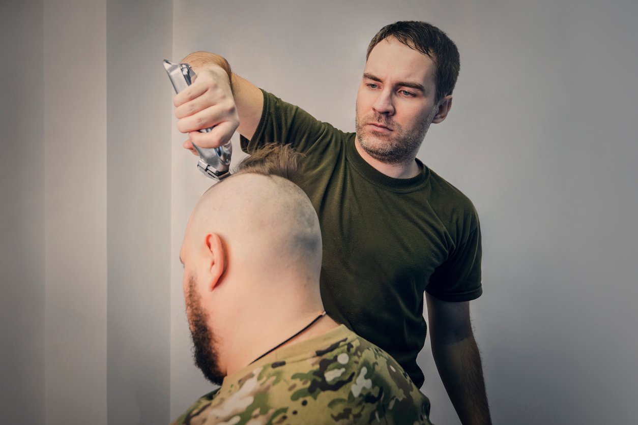 A man in a green shirt uses electric clippers to shave the head of another man wearing a camouflage shirt. The background is plain and neutral.
