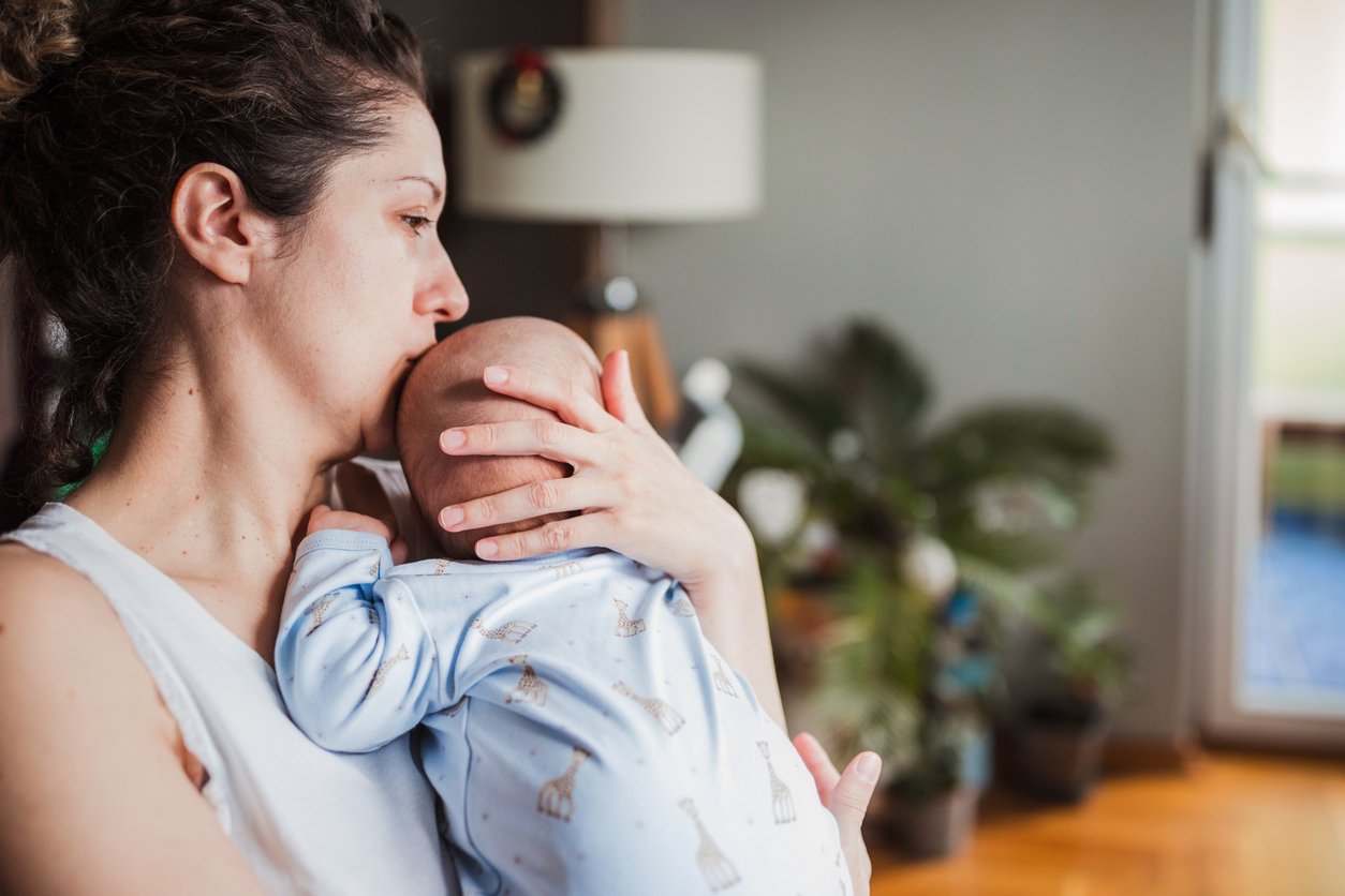 A woman sits indoors, gently holding and comforting a baby on her shoulder. She kisses the baby’s head, and both appear calm. A plant and a lamp are visible in the softly lit background.