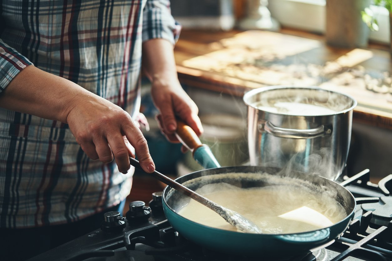 A person in a plaid shirt stirs a creamy sauce in a blue pan on a stove, with steam rising. A shiny pot with a lid is boiling on another burner in a sunlit kitchen.