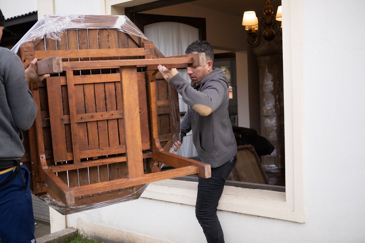 Two people are carefully carrying a large wooden table through a window opening, with one person inside and one outside a white building. The table is wrapped in plastic for protection.