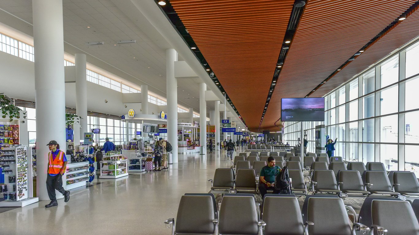 An airport terminal with empty gray seats, a few people waiting, large floor-to-ceiling windows, shops on the left, and a worker in an orange safety vest walking through the spacious area.