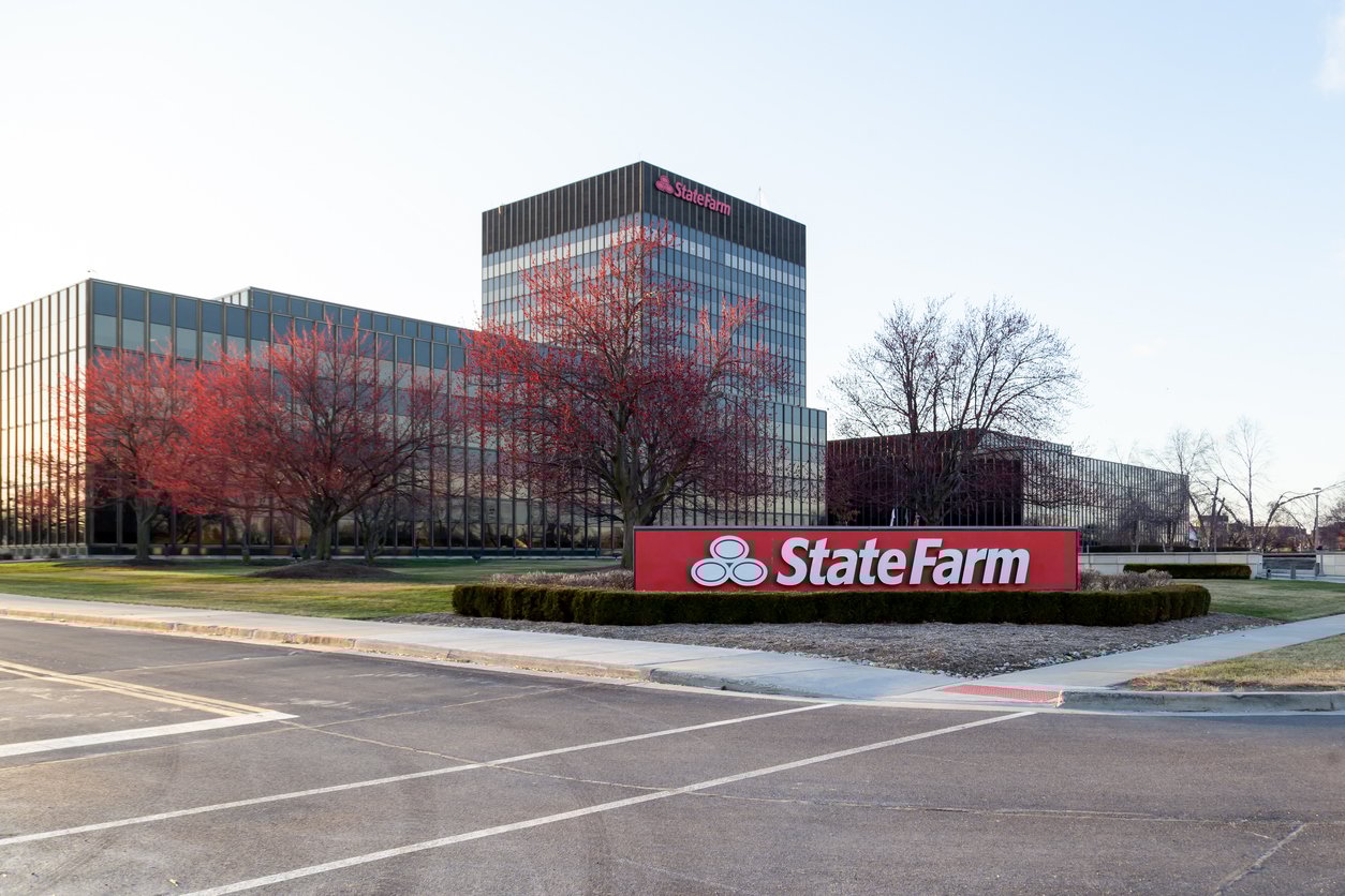 A large office building with reflective windows and red-leaved trees in front, featuring a prominent State Farm sign near the entrance on a landscaped island.