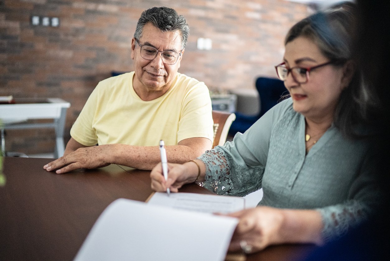 An older man in a yellow shirt sits at a table, watching as a woman in glasses and a light blue blouse writes on a document with a pen. They appear to be in a casual indoor setting.
