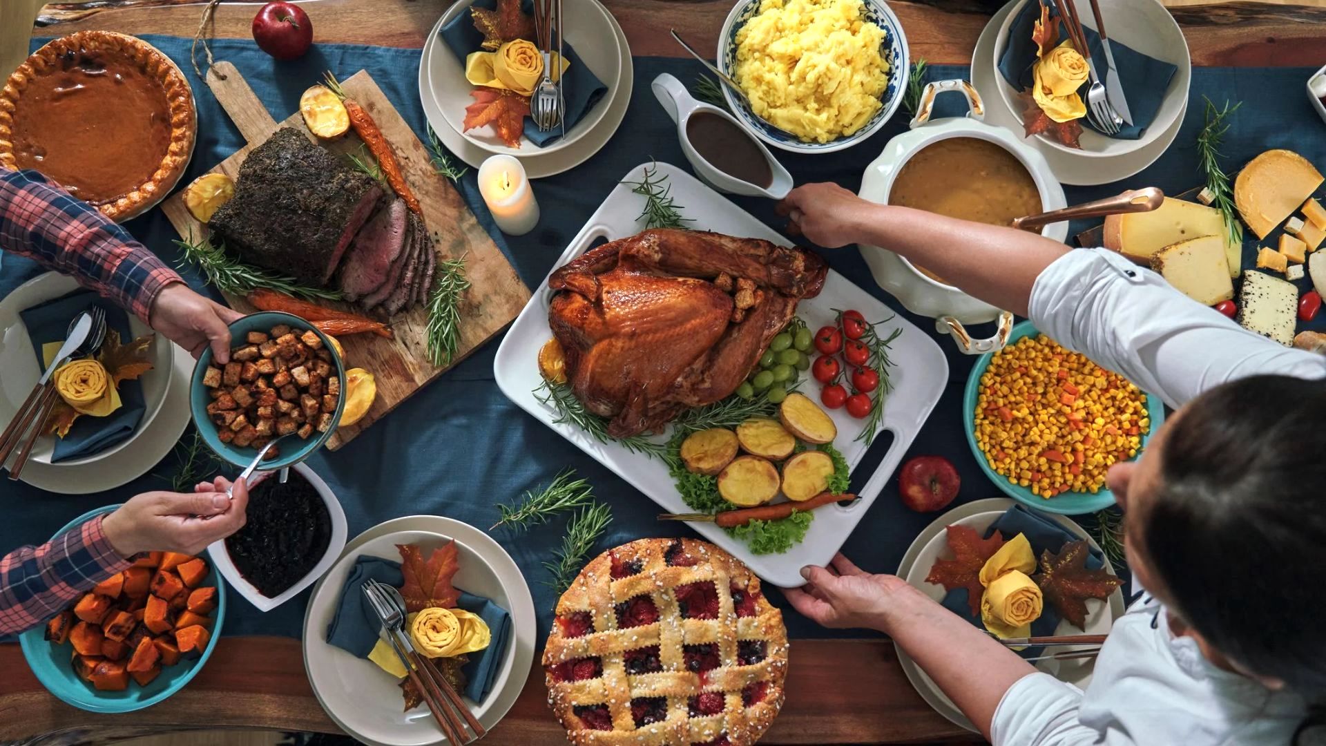 A table set for a festive meal with roast turkey, mashed potatoes, gravy, pie, vegetables, bread, and various side dishes. People are serving themselves and reaching for food.