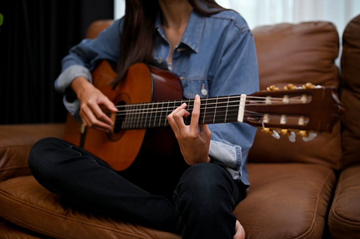 A person wearing a denim shirt and black pants sits cross-legged on a brown couch, playing an acoustic guitar. Their face is not visible, and their hands are positioned on the guitar’s neck and body.