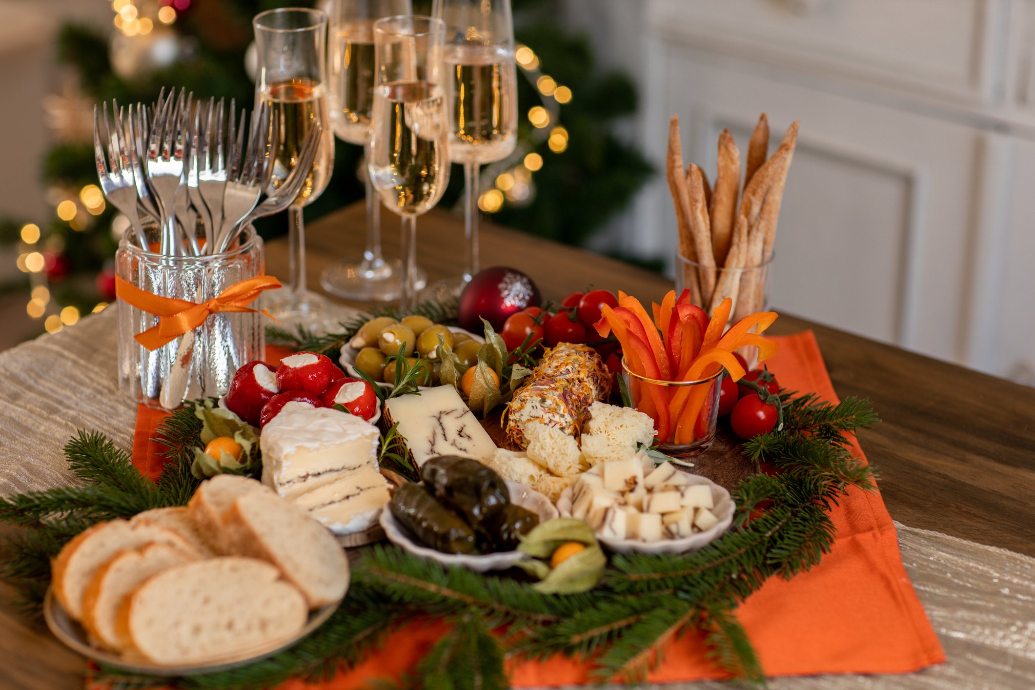 A festive holiday table displays assorted cheeses, bread slices, breadsticks, stuffed grape leaves, olives, vegetables, and four glasses of sparkling wine, decorated with greenery and orange accents.