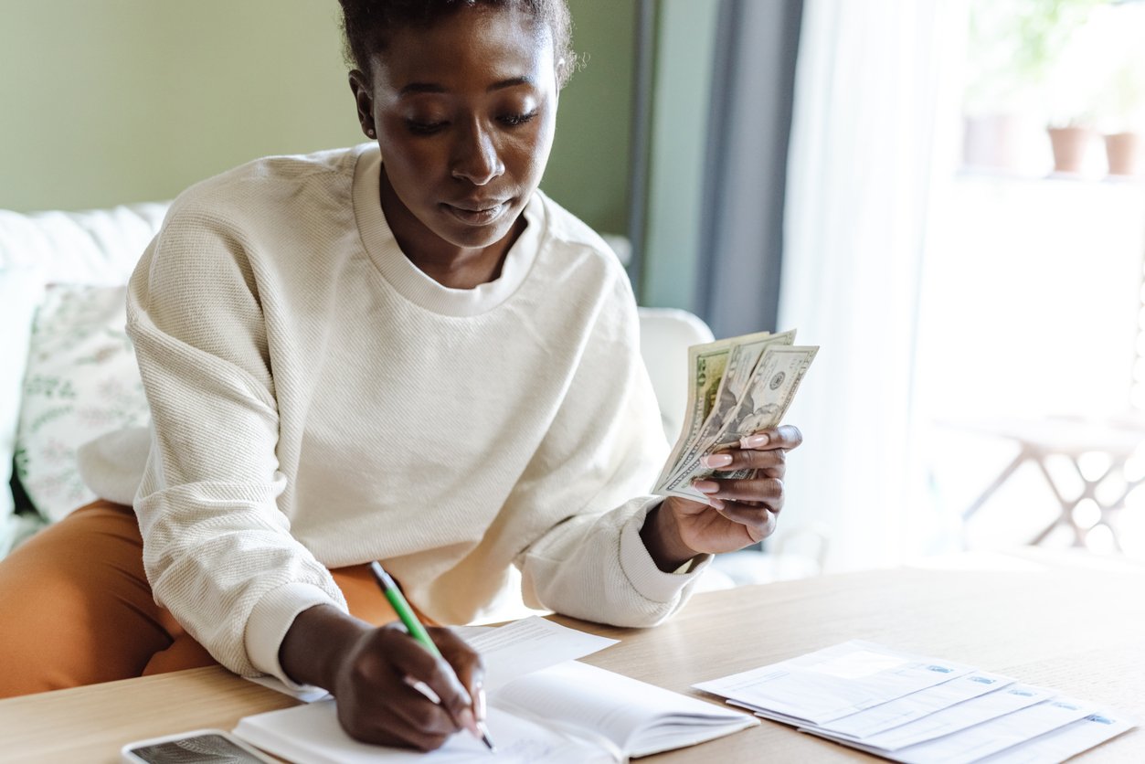 A woman sits at a table holding cash in one hand and writing in a notebook with the other, with documents and envelopes on the table, appearing to manage her finances.