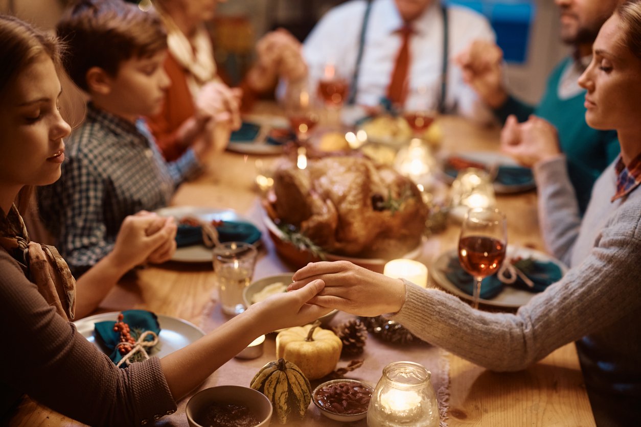 A family sits around a dinner table holding hands in prayer, with a roast turkey, candles, and autumn decorations in the center—capturing the warmth of a holiday gathering and evoking reflections on Thanksgiving dinner cost through the years.