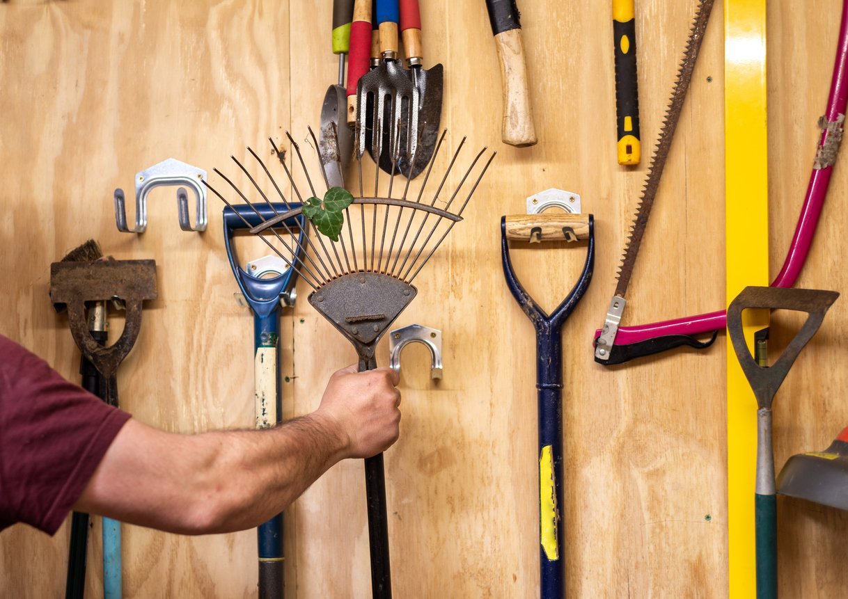 A person’s arm hangs a metal rake on a hook, surrounded by various garden tools neatly organized on a wooden wall.