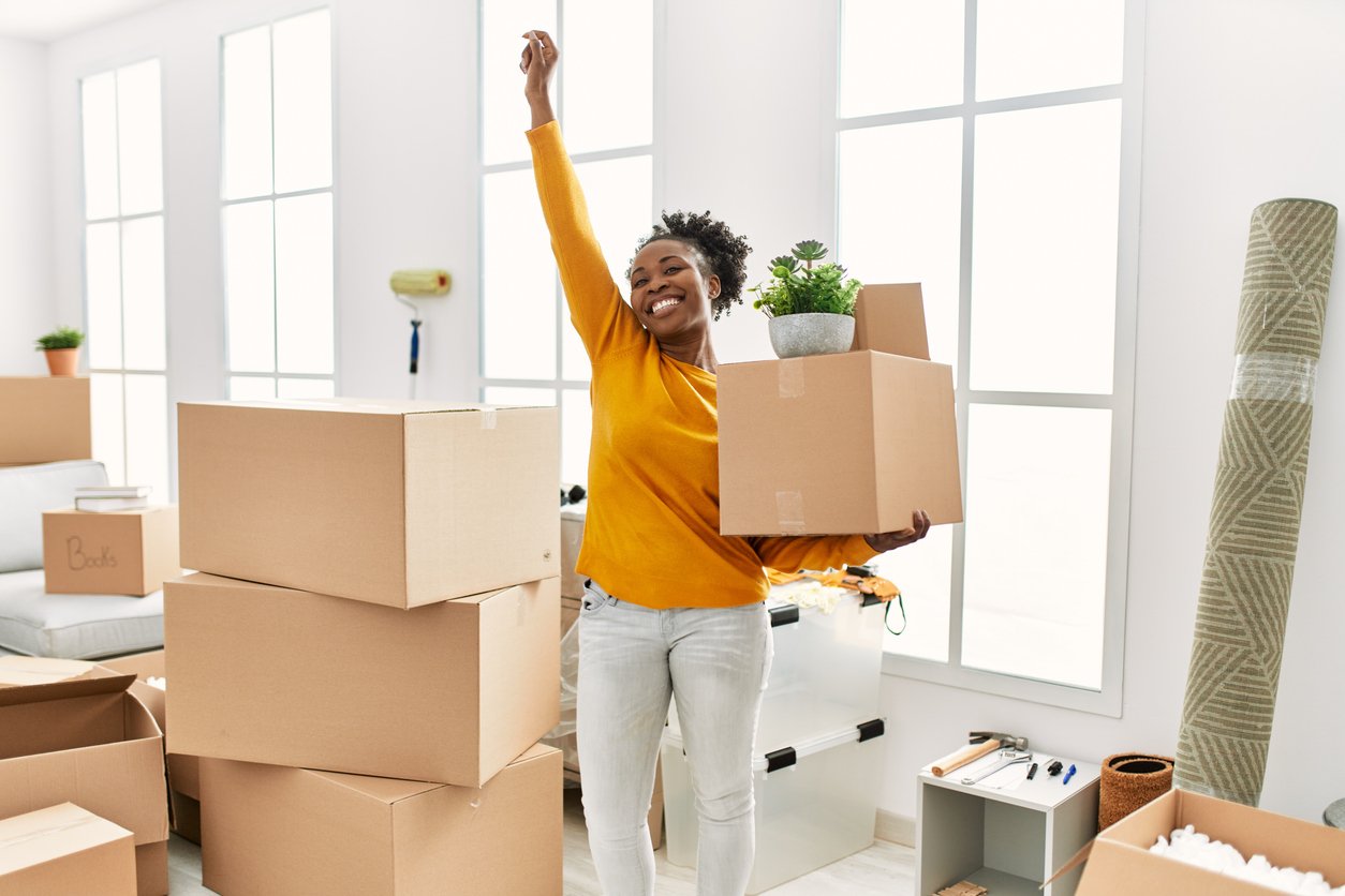 A smiling woman wearing a yellow sweater holds a box with a potted plant and raises one arm in celebration while standing among packed moving boxes in a bright room.