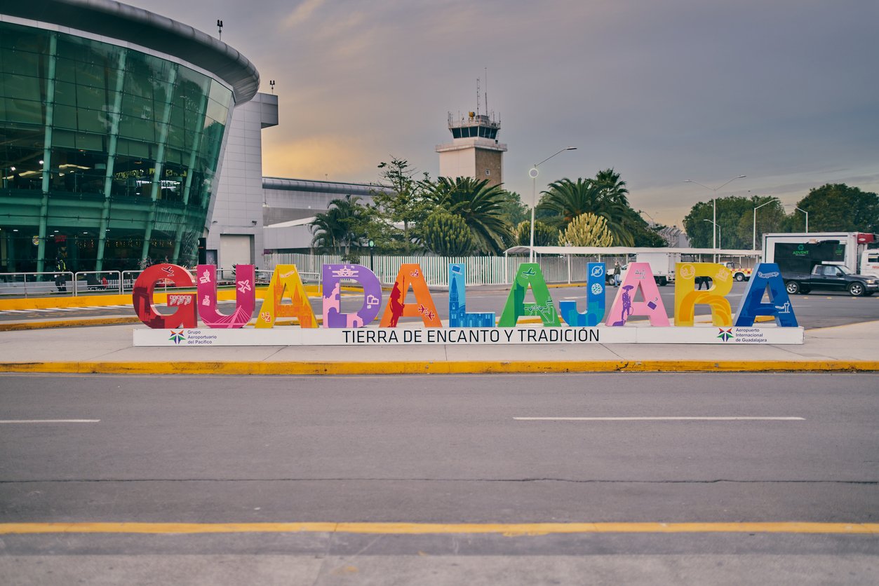 Colorful “Guadalajara” sign with the phrase “Tierra de encanto y tradición” stands outside a modern building near a street, with trees and a control tower visible in the background under a cloudy sky.