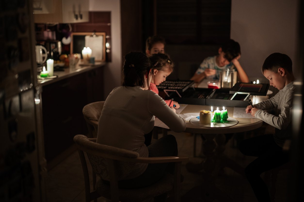A family sits around a table in a dimly lit room, doing homework and reading by candlelight during a power outage. The kitchen is visible in the background with more candles providing soft light.
