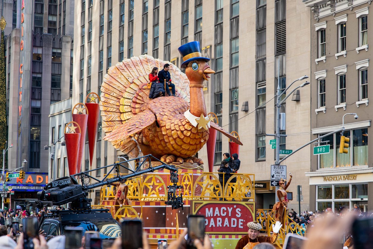 A giant turkey float with people riding on top moves through a city street during the Macy's Thanksgiving Day Parade, reminding spectators of Thanksgiving Dinner Cost History amid the festive atmosphere, buildings, cameras, and cheerful crowds.