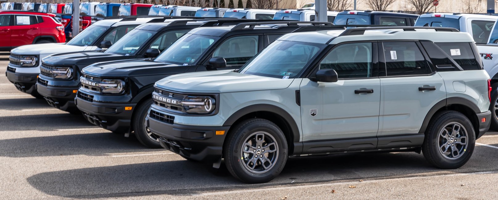 Four new Ford Bronco Sport SUVs, two black and two light gray, are parked in a row at a car dealership lot under daylight, with other vehicles and buildings visible in the background.