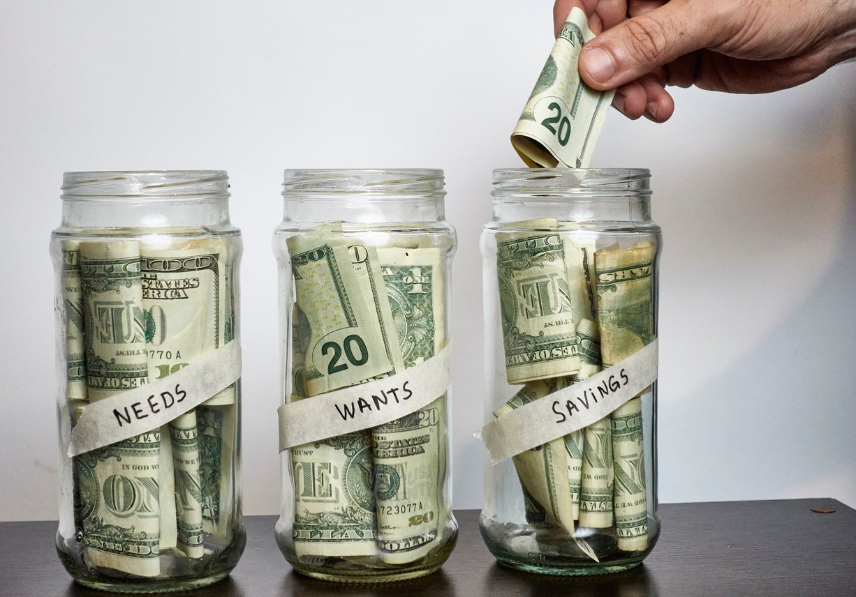 Three glass jars labeled "Needs," "Wants," and "Savings," each containing U.S. dollar bills. A hand places a $20 bill into the "Savings" jar. The jars are on a wooden surface against a plain background.